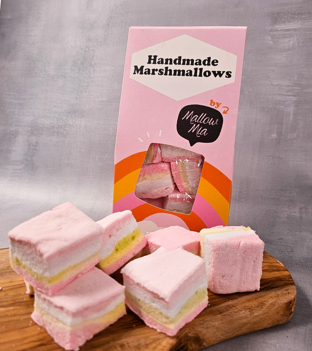 Box of Mallow Mia handmade rainbow marshmallows with pastel pink, white, and yellow layers, stacked on a wooden board in front of bright pink packaging with orange and yellow stripes.