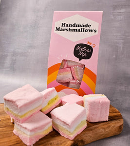 Box of Mallow Mia handmade rainbow marshmallows with pastel pink, white, and yellow layers, stacked on a wooden board in front of bright pink packaging with orange and yellow stripes.