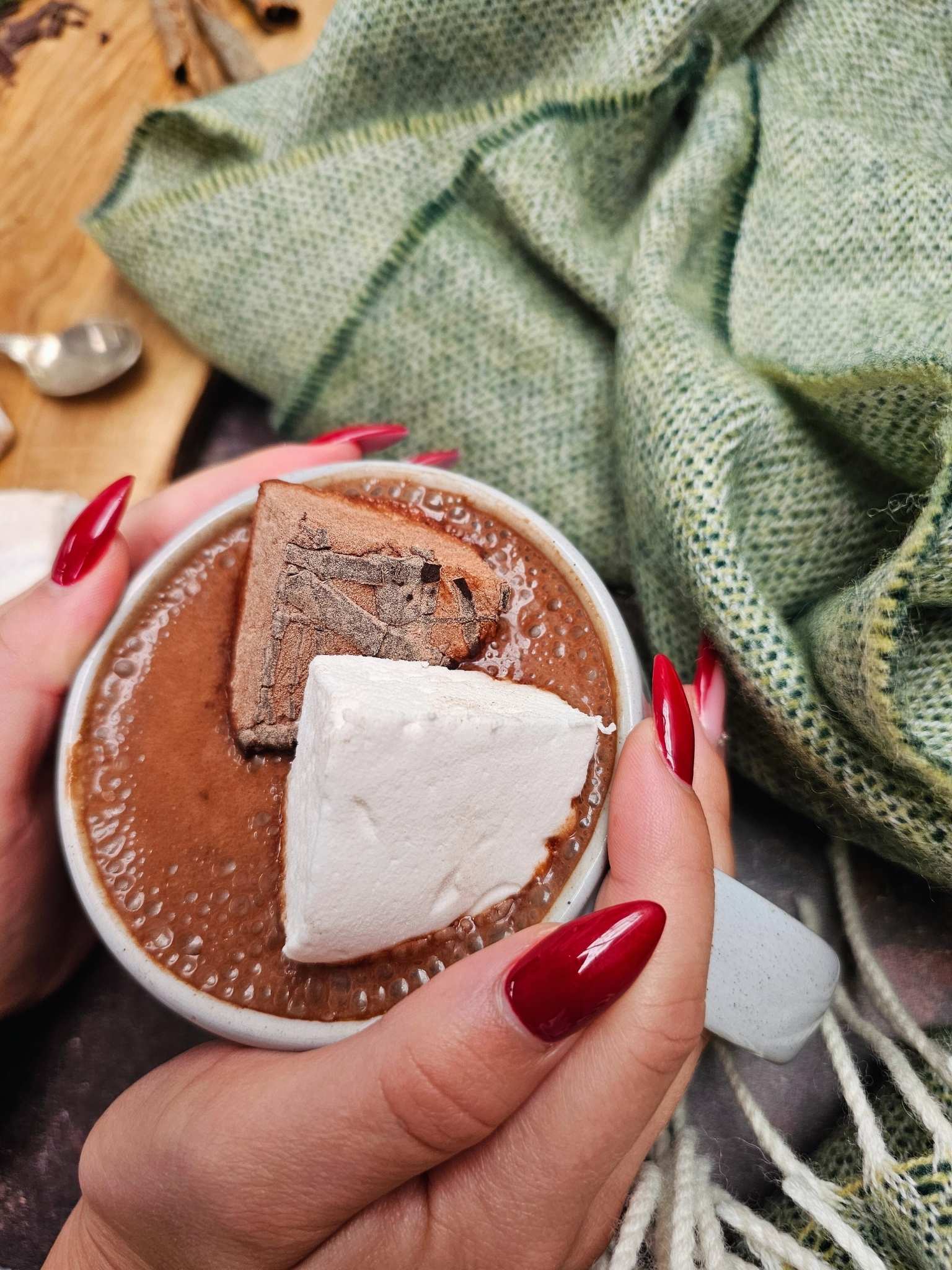 Hot chocolate with a marshmallow in a white cup, held by a person with red nails, on a wooden surface with a green blanket.
