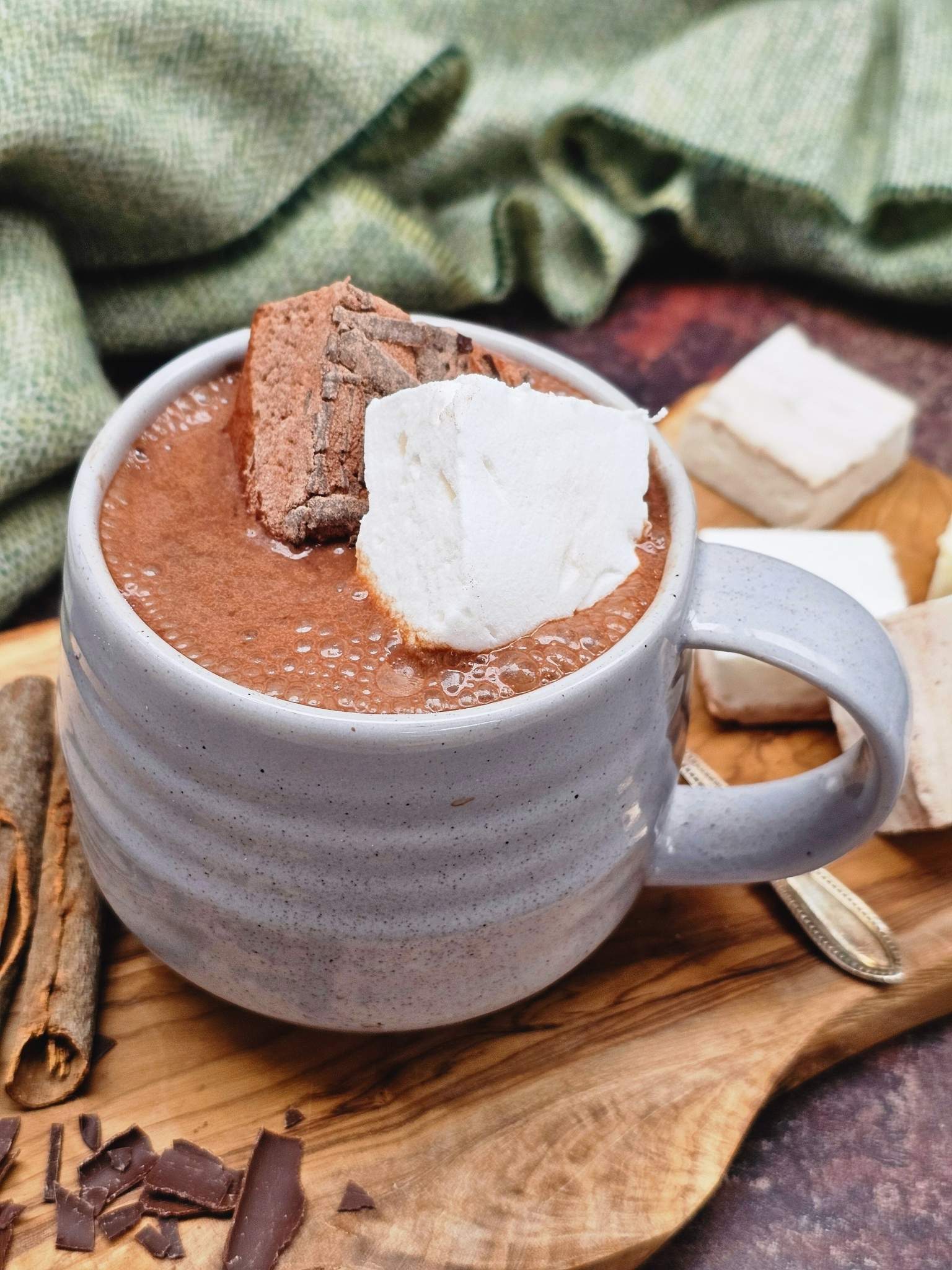 Hot chocolate in a white mug with marshmallows, chocolate pieces, and cinnamon sticks on a wooden surface.