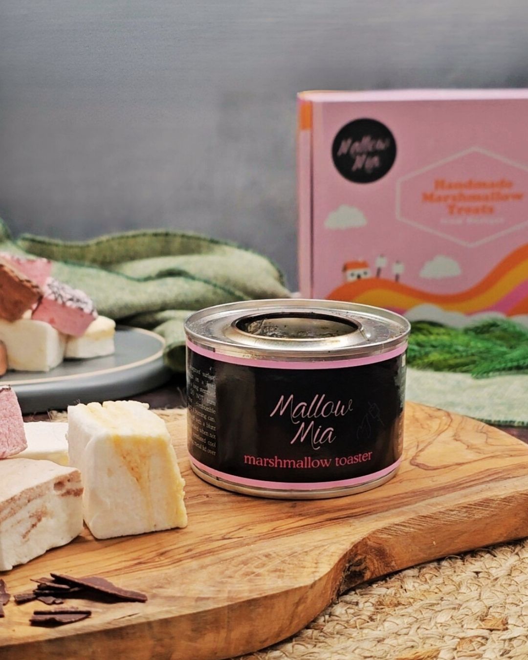 Marshmallow toaster canister with 'Mallow Mia' branding on a wooden board with marshmallows and a pink box in the background.