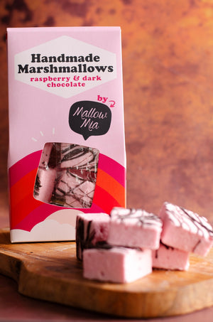 Box of Mallow Mia handmade raspberry and dark chocolate marshmallows with pink packaging, shown with several fluffy pink marshmallows drizzled in dark chocolate on a wooden board.