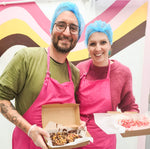 Two smiling adults wearing pink aprons and blue hairnets holding trays of decorated marshmallow treats at a Mallow Mia workshop, standing in front of a colourful striped wall.